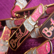 Load image into Gallery viewer, A collection of gold enamel pins of a open black tube of pink glittery lipstick with &quot;Butch&quot; in pink glitter is written on the side of the tube on top of fuchsia fabric